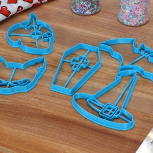 May include: A set of five blue plastic Halloween cookie cutters. The cutters are shaped like a pumpkin, a cat, a coffin, a witch's hat, and a bat.