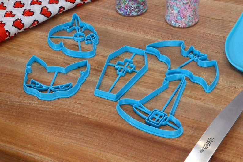 May include: A set of five blue plastic Halloween cookie cutters. The cutters are shaped like a pumpkin, a cat, a coffin, a witch's hat, and a bat.