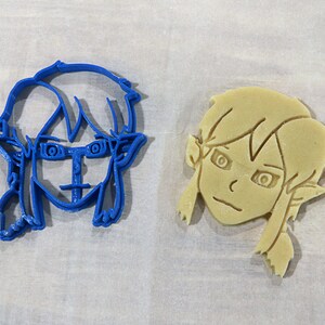 May include: Two cookie cutters, one blue and one beige, shaped like a cartoon character with large eyes and elf ears.