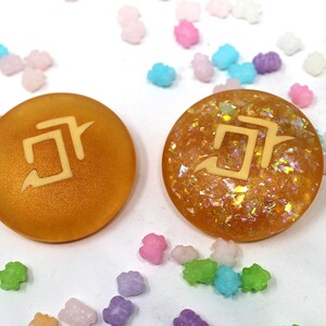May include: Two round, gold-colored resin cabochons with a white symbol on each. The cabochon on the left is matte and the cabochon on the right is glittery.