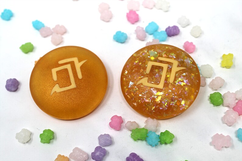 May include: Two round, gold-colored resin cabochons with a white symbol on each. The cabochon on the left is matte and the cabochon on the right is glittery.