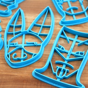 May include: A set of blue plastic cookie cutters shaped like a bunny, a face, and a glass. The cutters are on a wooden surface.