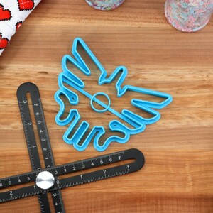 May include: A blue plastic cookie cutter shaped like a stylized bird with a hole in the center. The cutter is on a wooden surface.