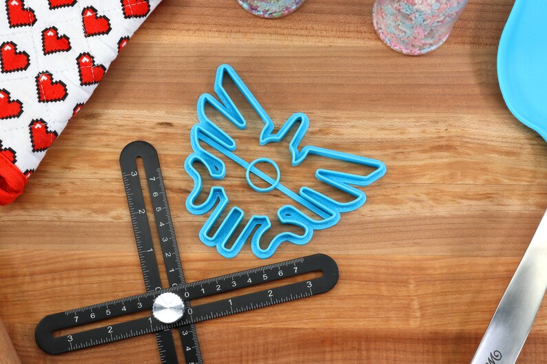 May include: A blue plastic cookie cutter shaped like a stylized bird with a hole in the center. The cutter is on a wooden surface.