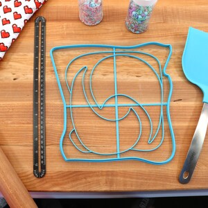 May include: A blue plastic bread mold shaped like a slice of bread with a swirl design on a wooden cutting board. There is a rolling pin, a ruler, a spatula, and two jars of sprinkles in the background.