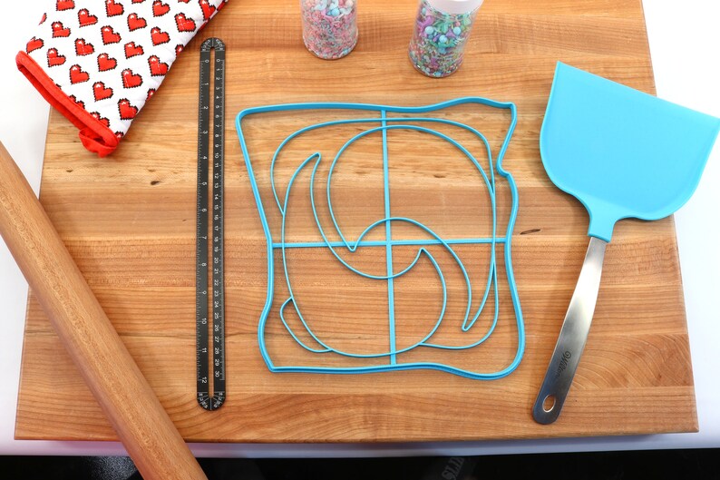 May include: A blue plastic bread mold shaped like a slice of bread with a swirl design on a wooden cutting board. There is a rolling pin, a ruler, a spatula, and two jars of sprinkles in the background.