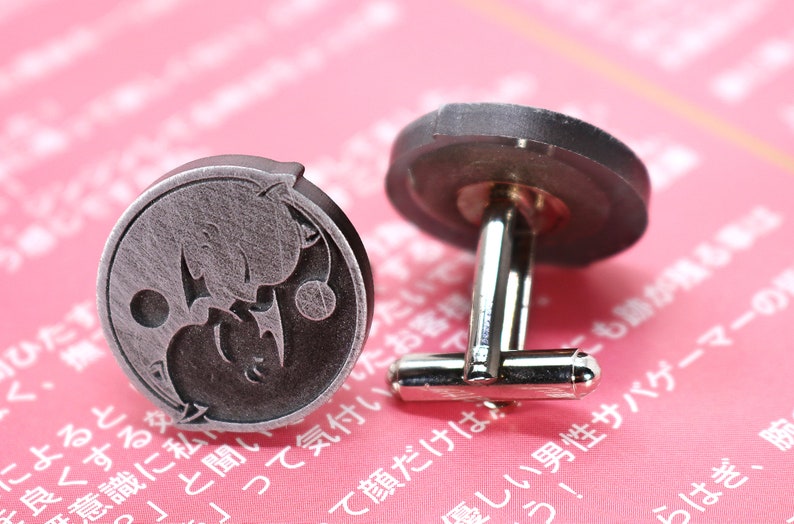May include: A pair of silver cufflinks with a yin yang design featuring two cats. The cufflinks are on a pink background.