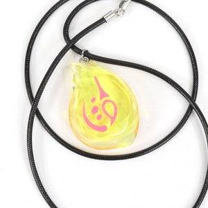 May include: A yellow teardrop-shaped pendant with a pink symbol inside, hanging from a black cord necklace.