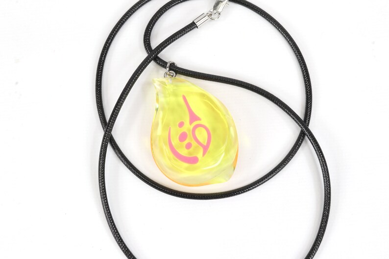 May include: A yellow teardrop-shaped pendant with a pink symbol inside, hanging from a black cord necklace.