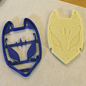 May include: A blue plastic fox-shaped cookie cutter next to a fox-shaped cookie on parchment paper. The cookie cutter has a detailed design, and the cookie shows the embossed fox face. The background includes a patterned fabric.