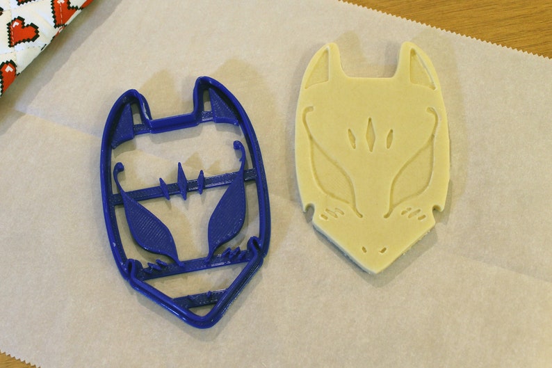 May include: A blue plastic fox-shaped cookie cutter next to a fox-shaped cookie on parchment paper. The cookie cutter has a detailed design, and the cookie shows the embossed fox face. The background includes a patterned fabric.