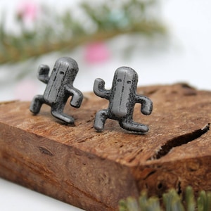 May include: A pair of silver-toned earrings featuring a stylized cactus character design. The earrings are displayed on a piece of wood.