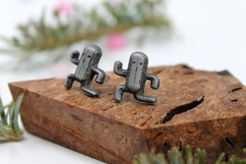 May include: A pair of silver-toned earrings featuring a stylized cactus character design. The earrings are displayed on a piece of wood.
