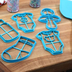 May include: A set of six blue plastic cookie cutters shaped like a baby bottle, two baby onesies, a sock, and a rattle. The cutters are on a wooden surface.