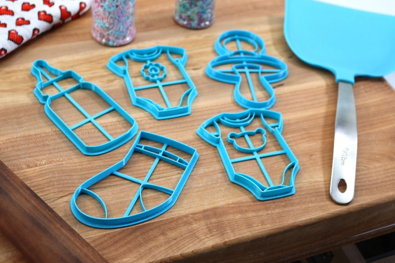 May include: A set of six blue plastic cookie cutters shaped like a baby bottle, two baby onesies, a sock, and a rattle. The cutters are on a wooden surface.
