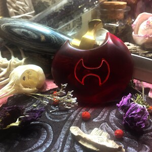 May include: A red glass bottle stopper with a gold top and a double-headed axe design. The bottle stopper is sitting on a dark surface with dried flowers and other decorative items.