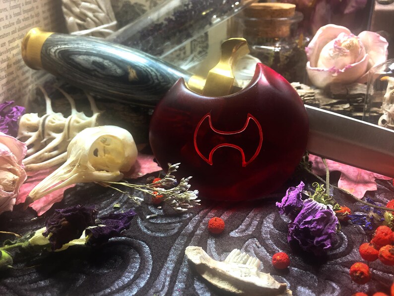 May include: A red glass bottle stopper with a gold top and a double-headed axe design. The bottle stopper is sitting on a dark surface with dried flowers and other decorative items.