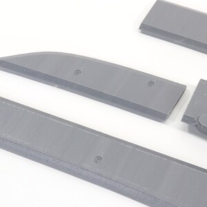 May include: Four gray plastic pieces, possibly parts of a model airplane or other model kit. The pieces are flat and rectangular with rounded edges. Some pieces have small holes.