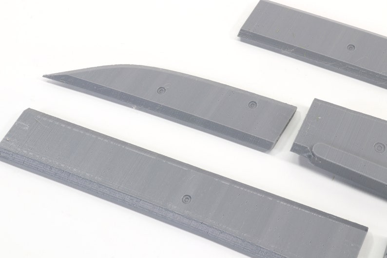 May include: Four gray plastic pieces, possibly parts of a model airplane or other model kit. The pieces are flat and rectangular with rounded edges. Some pieces have small holes.
