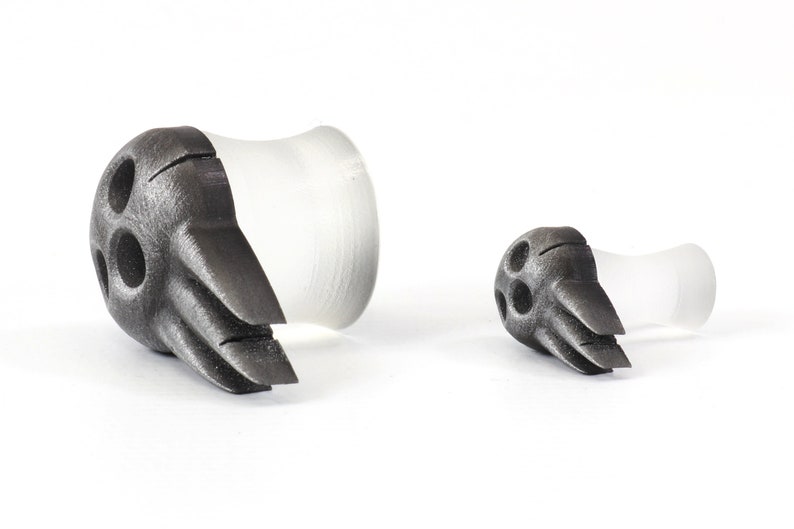 May include: Two ear gauges, one large and one small, with a dark gray, textured, bird-like design on the front. The back portion of the gauges is clear and cylindrical. The gauges are displayed on a white background.