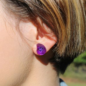 May include: A close-up of a person's ear with a purple ear gauge. The ear gauge is shaped like a teardrop and has a small white design on it.