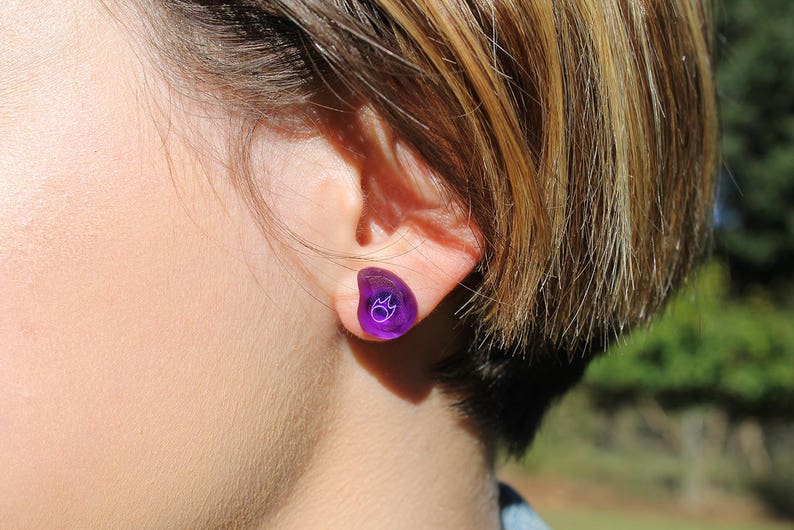 May include: A close-up of a person's ear with a purple ear gauge. The ear gauge is shaped like a teardrop and has a small white design on it.