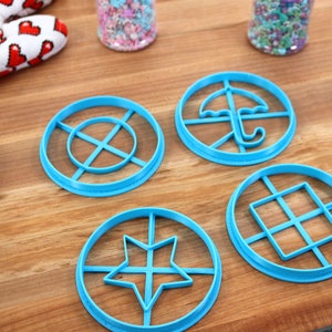 May include: Four light blue cookie cutters with different shapes: a star, an umbrella, a square grid, and a circle with lines. The cutters are on a wooden surface, with a red and white heart-patterned oven mitt and sprinkles in the background.