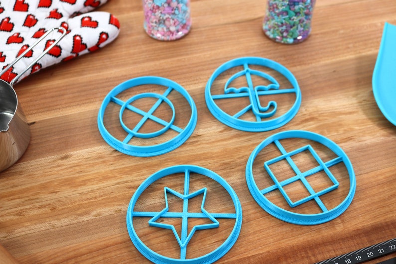 May include: Four light blue cookie cutters with different shapes: a star, an umbrella, a square grid, and a circle with lines. The cutters are on a wooden surface, with a red and white heart-patterned oven mitt and sprinkles in the background.
