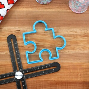May include: A blue plastic puzzle-shaped cookie cutter on a wooden surface with a black measuring tool.