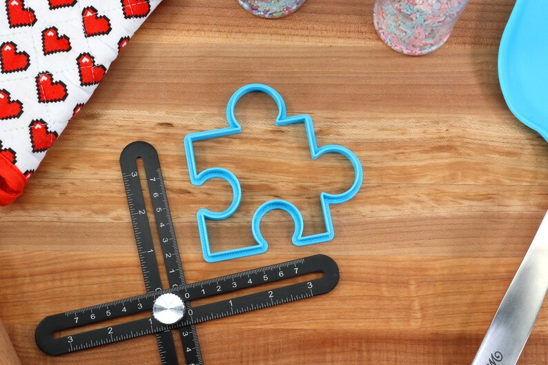 May include: A blue plastic puzzle-shaped cookie cutter on a wooden surface with a black measuring tool.