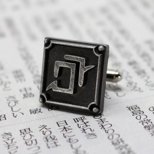 K&ouml;nnte beinhalten: Ein quadratischer, dunkelgrauer Manschettenknopf aus Metall mit einem stilisierten Design. Der Manschettenknopf befindet sich vor einem verschwommenen Hintergrund mit japanischem Text. Der Manschettenknopf hat einen silberfarbenen Stift.