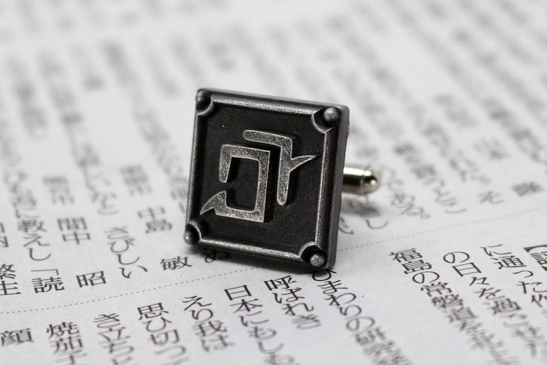 K&ouml;nnte beinhalten: Ein quadratischer, dunkelgrauer Manschettenknopf aus Metall mit einem stilisierten Design. Der Manschettenknopf befindet sich vor einem verschwommenen Hintergrund mit japanischem Text. Der Manschettenknopf hat einen silberfarbenen Stift.