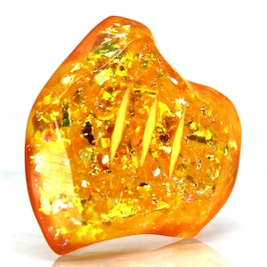 May include: A large, irregular shaped piece of amber with a golden yellow color and a translucent appearance. The amber has a unique pattern of inclusions that resemble small, shimmering flakes.