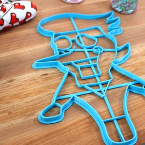 May include: A blue plastic cookie cutter shaped like a cartoon character with a hat, glasses, and a mustache. The cutter is on a wooden surface.