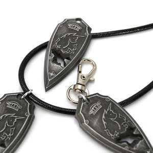 May include: Three silver-toned pendants with a unicorn design, hanging on a black cord. The pendants are shaped like shields with a crown on top.
