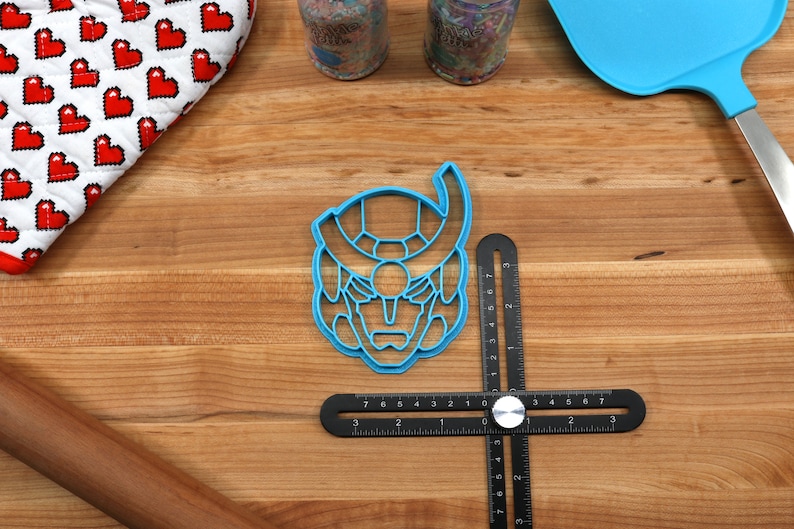 May include: Blue plastic cookie cutter shaped like a robot head with a detailed design. The cutter is on a wooden surface with a ruler and a rolling pin.