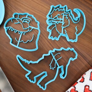 May include: Three blue plastic dinosaur cookie cutters on a wooden surface. The cutters are shaped like a dinosaur head, a standing dinosaur, and a dinosaur with its head down.