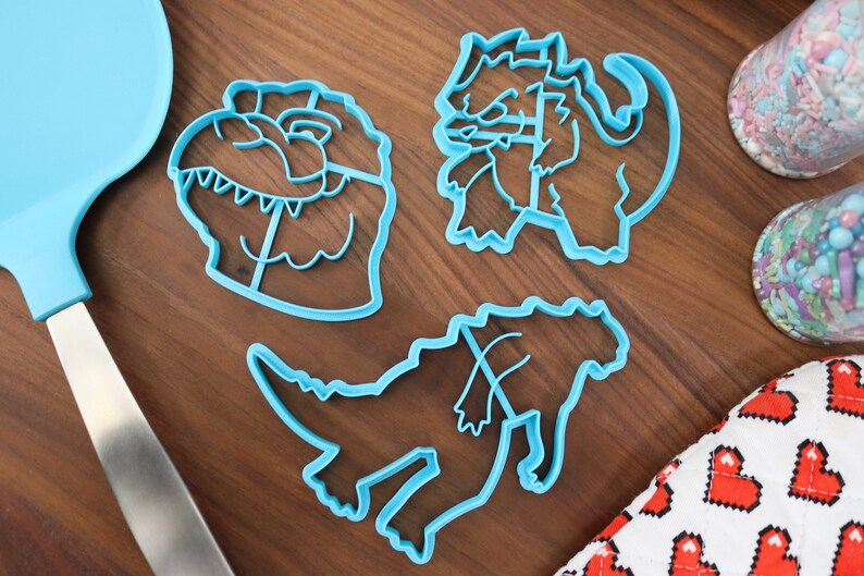 May include: Three blue plastic dinosaur cookie cutters on a wooden surface. The cutters are shaped like a dinosaur head, a standing dinosaur, and a dinosaur with its head down.