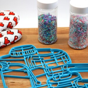 May include: Blue plastic cookie cutter shaped like a snowman with a hat and scarf. Two jars of colorful sprinkles are in the background.