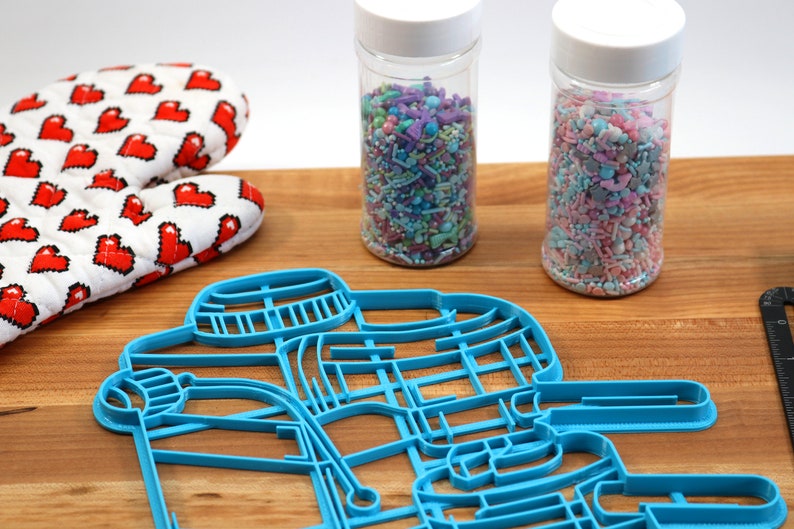 May include: Blue plastic cookie cutter shaped like a snowman with a hat and scarf. Two jars of colorful sprinkles are in the background.