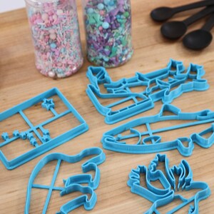 May include: A set of blue plastic cookie cutters shaped like a fish, a Christmas tree, a reindeer, a star, and a hand. The cutters are on a wooden surface with two jars of sprinkles in the background.