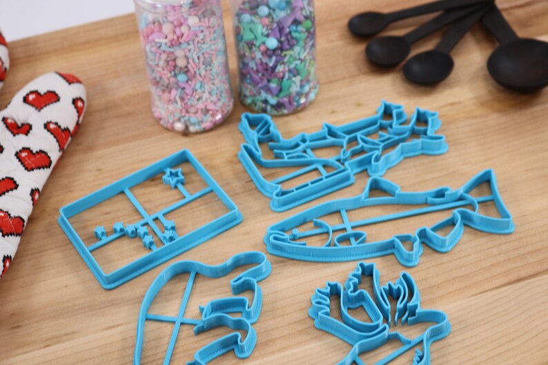 May include: A set of blue plastic cookie cutters shaped like a fish, a Christmas tree, a reindeer, a star, and a hand. The cutters are on a wooden surface with two jars of sprinkles in the background.