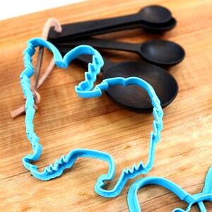 May include: Two blue plastic cookie cutters shaped like a dog and a heart on a wooden surface. A set of black measuring spoons are also in the image.