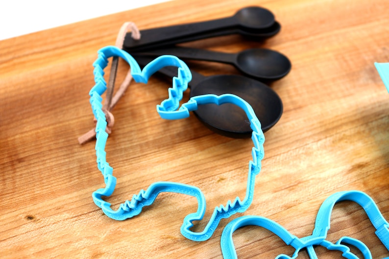 May include: Two blue plastic cookie cutters shaped like a dog and a heart on a wooden surface. A set of black measuring spoons are also in the image.