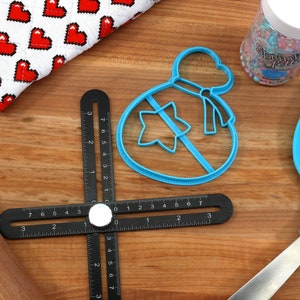 May include: Blue plastic cookie cutter shaped like a bag with a star inside. The cutter is on a wooden surface with a black ruler, a rolling pin, a blue spatula, and a jar of sprinkles.