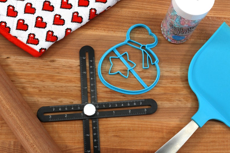 May include: Blue plastic cookie cutter shaped like a bag with a star inside. The cutter is on a wooden surface with a black ruler, a rolling pin, a blue spatula, and a jar of sprinkles.