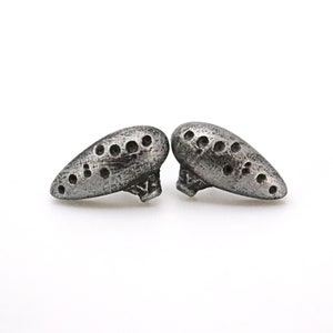 Pu&ograve; includere: Due orecchini color argento a forma di ocarina. Ogni orecchino presenta una serie di piccoli fori distanziati e una superficie strutturata. Sono esposti su sfondo bianco.