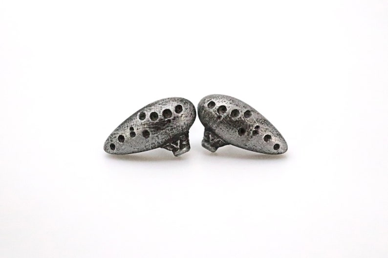 Pu&ograve; includere: Due orecchini color argento a forma di ocarina. Ogni orecchino presenta una serie di piccoli fori distanziati e una superficie strutturata. Sono esposti su sfondo bianco.