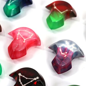 May include: A set of nine colourful resin dice with constellations embedded inside. Each die is shaped like a crescent moon and features a different constellation.