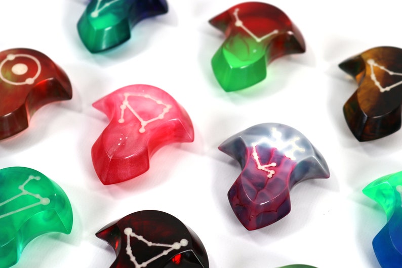 May include: A set of nine colourful resin dice with constellations embedded inside. Each die is shaped like a crescent moon and features a different constellation.